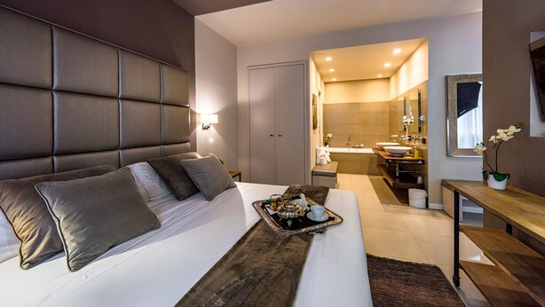 Ein modernes Hotelzimmer mit einem großen Bett, einem Nachttisch, einem Tablett mit Frühstücksangeboten und einem Badezimmer mit Waschbecken und Spiegel.