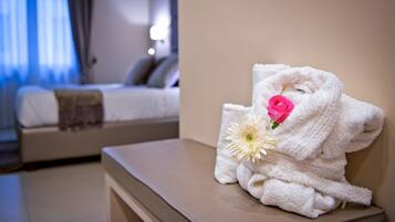 A hotel room with a neatly made bed, a bedside table with a towel and a flower, and a view of another room in the background.