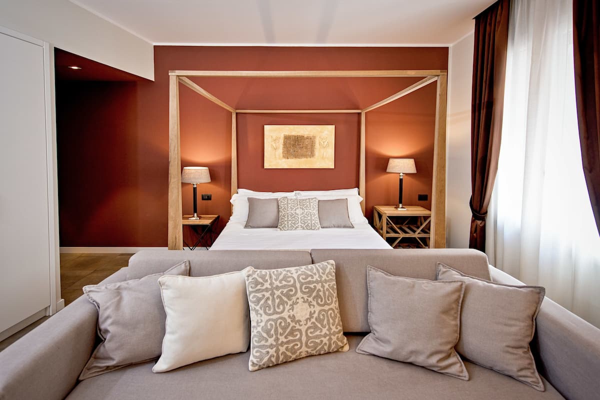 Une chambre d’hôtel avec un lit à baldaquin, un canapé agrémenté de coussins à motifs, deux tables de chevet avec des lampes et une œuvre d’art encadrée au mur.