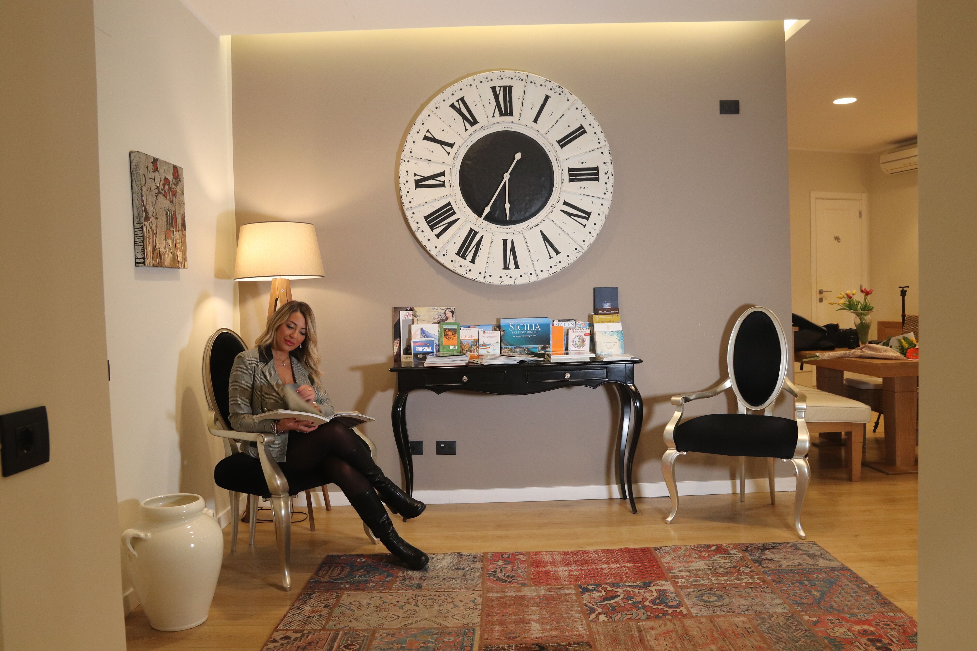 Una habitación con un reloj de pared grande, un escritorio con libros, una silla y una alfombra estampada.