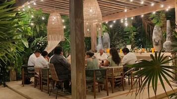 Un área de comedor al aire libre con mesas y sillas de madera, rodeada de abundante follaje y lámparas colgantes.