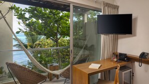 A room with a wooden desk, a television, a telephone, and a small refrigerator. There is a hammock chair and a view of the ocean through a glass door. - Mereva Tulum (Tulum)