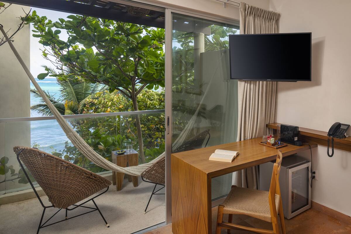 A room with a wooden desk, a television, a telephone, and a small refrigerator. There is a hammock chair and a view of the ocean through a glass door.