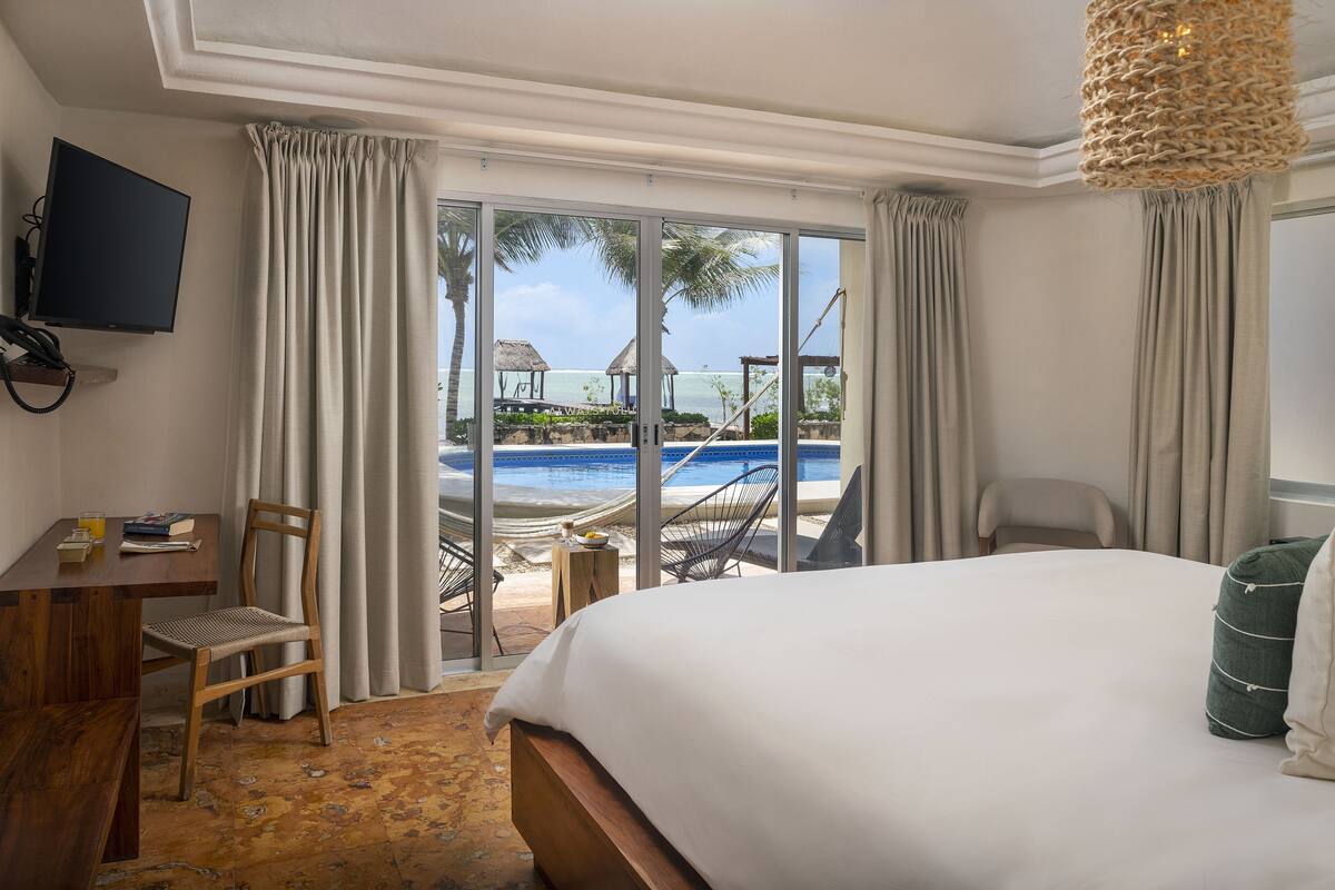 A hotel room with a bed, a desk, a chair, and a TV. There is a view of a beach and a pool outside the window.