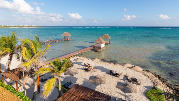 A beach with thatched huts, palm trees, and a wooden pier extending into the sea. - Mereva Tulum (Tulum)