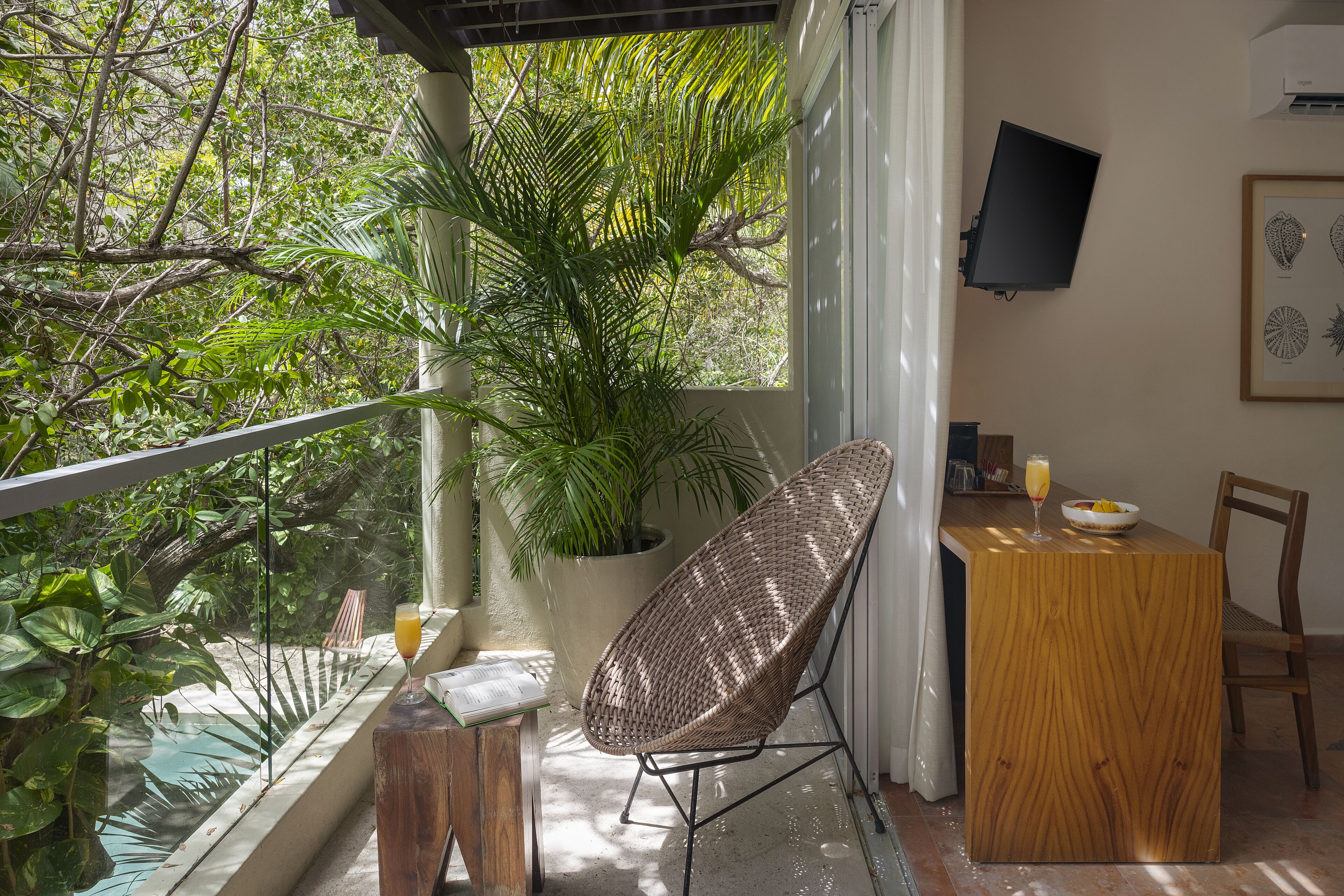 Un balcon avec un fauteuil en osier, une table en bois, une plante en pot et un téléviseur fixé au mur.
