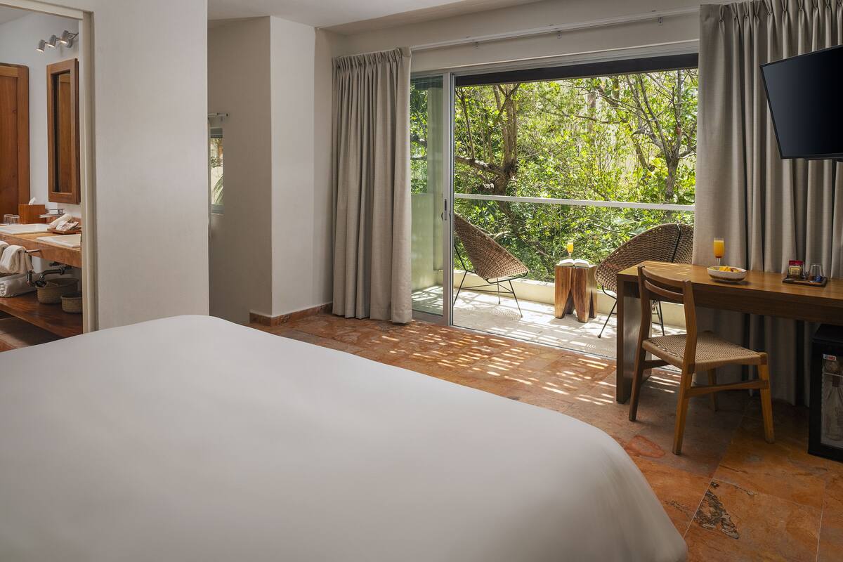 A hotel room with a large bed, a desk with a chair, a TV, and a balcony with a view of trees.