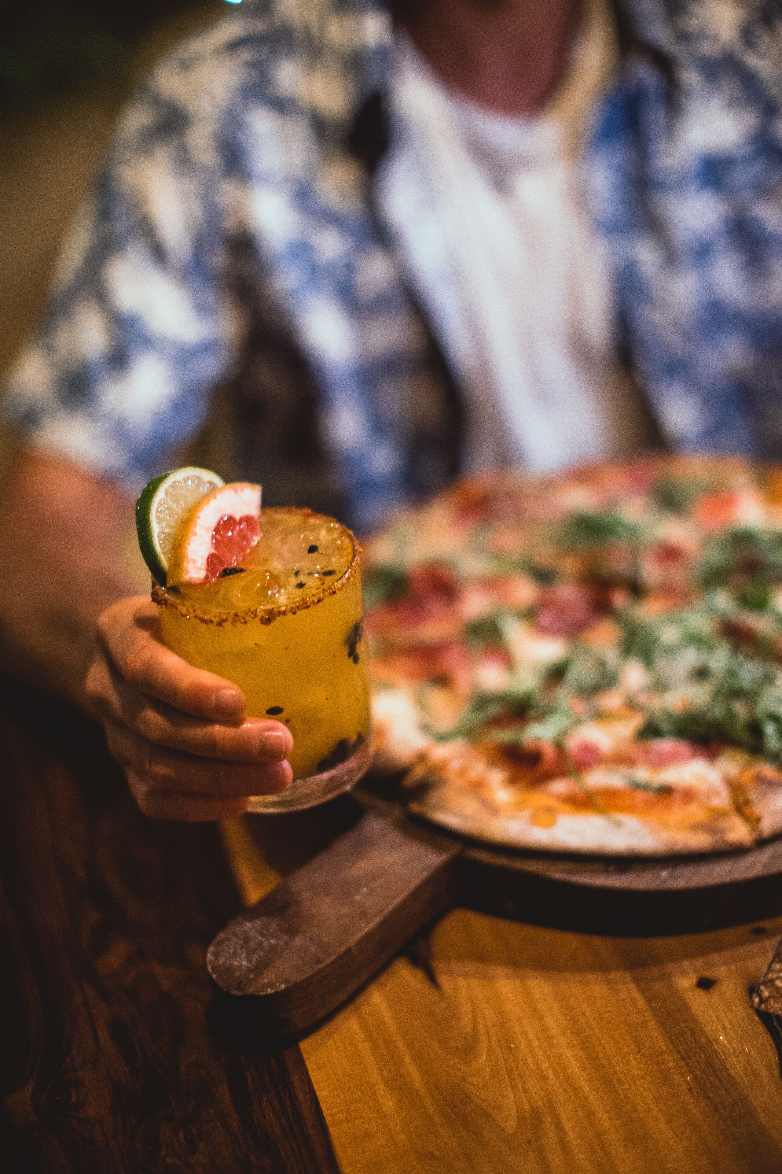 A person holding a cocktail with a lime garnish, a pizza on a wooden board, and a wooden table.