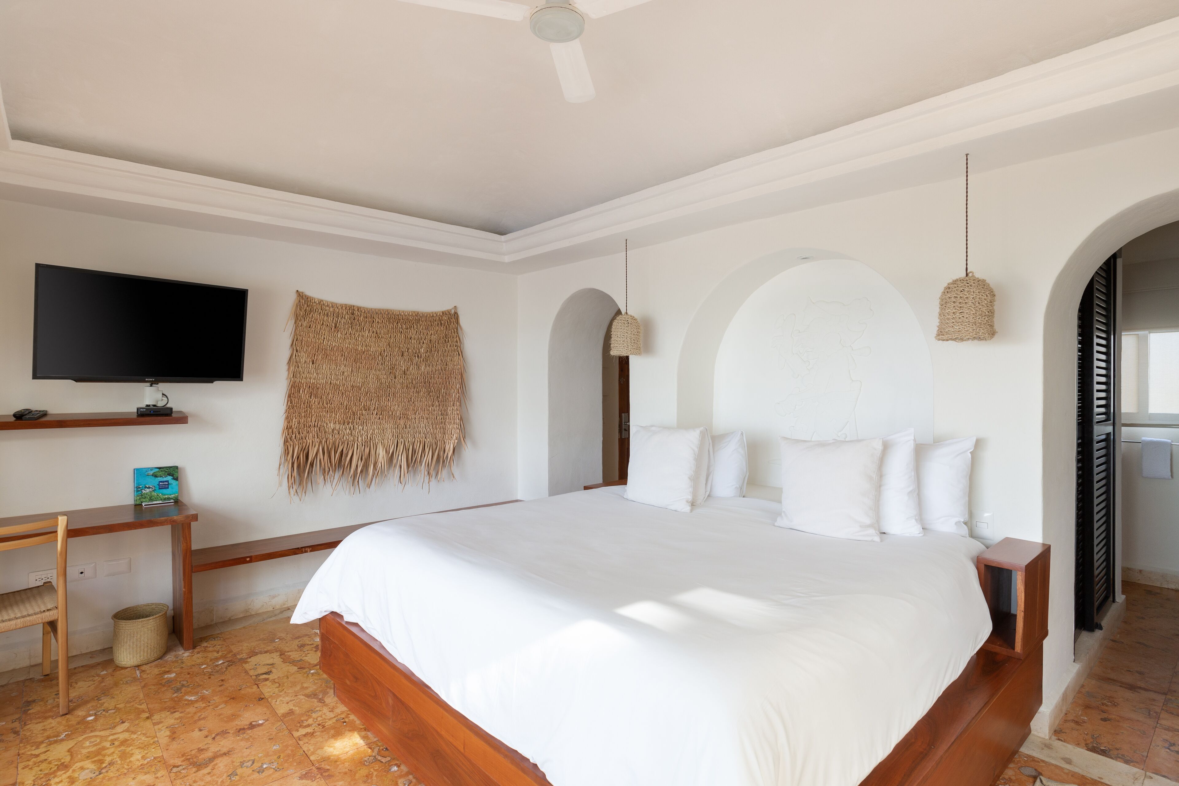 A bedroom with a large bed, a TV on a shelf, a woven wall hanging, and a ceiling fan.