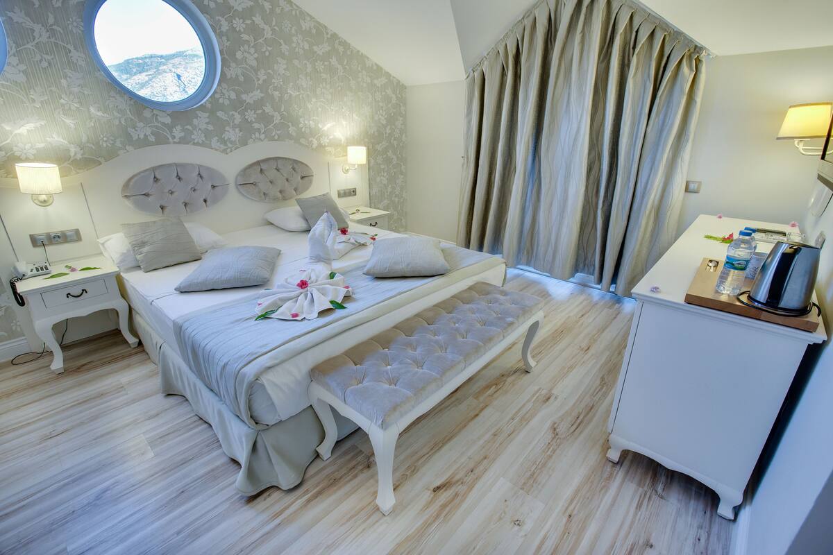 A bedroom with a bed, bench, bedside tables, a round window, curtains, and a desk with a lamp.