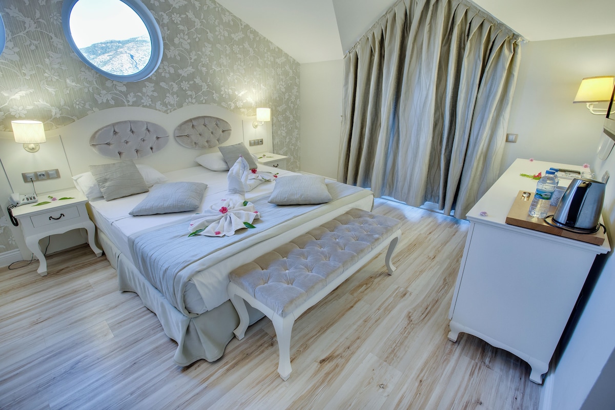 A bedroom with a large bed, a bench, a bedside table, a lamp, and a view of the outdoors.