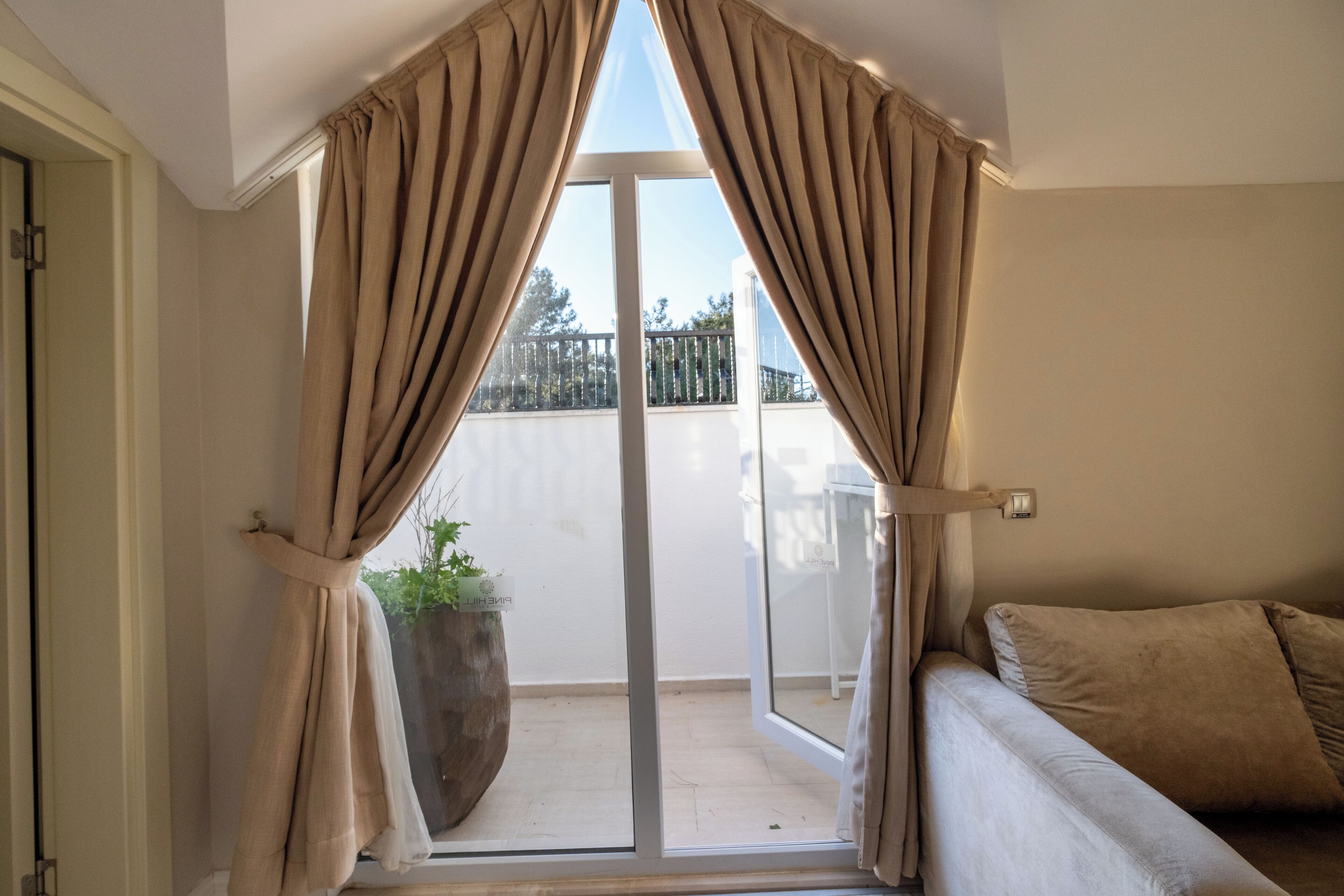 A room with a sliding glass door leading to a balcony, beige curtains, a potted plant, and a beige sofa.