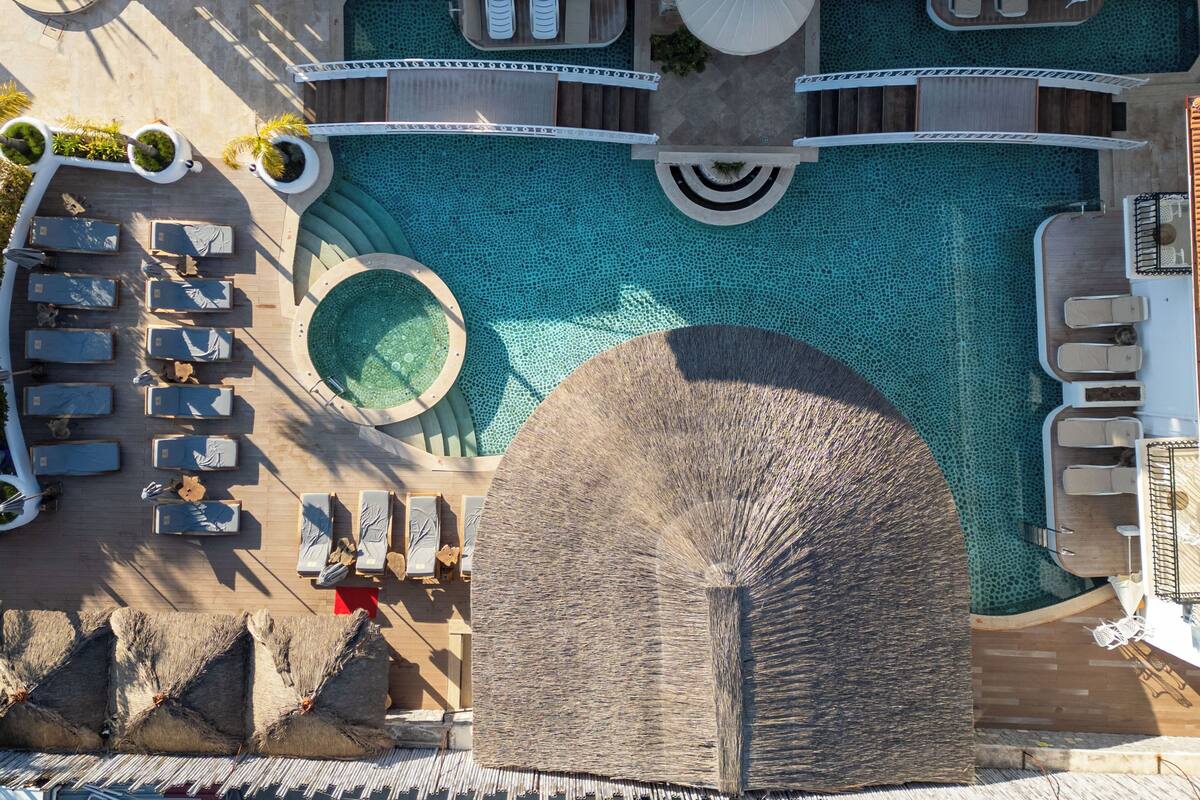 An aerial view of a resort's pool area with sun loungers, a central circular hot tub, and a stone structure.