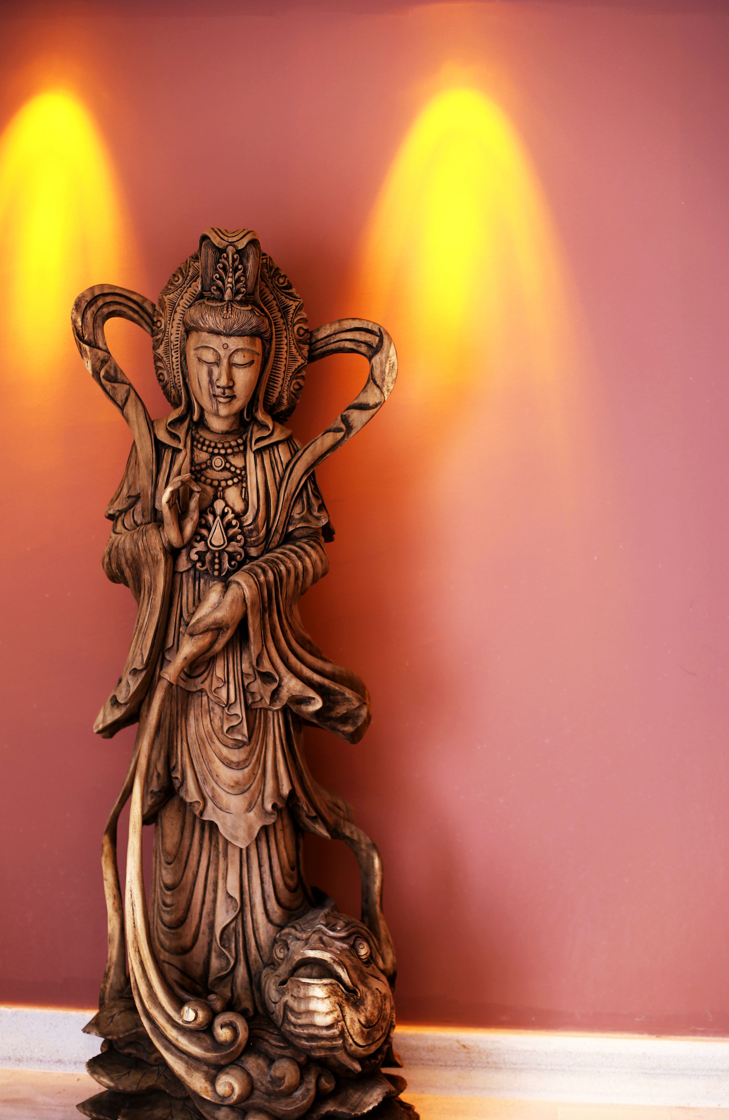 A statue of a figure with elaborate attire and a headpiece, standing against a red background with two light sources.