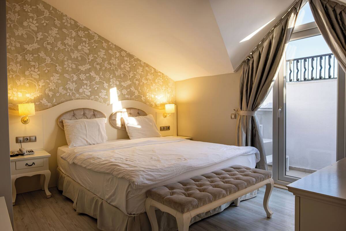 A bedroom with a large bed, bedside lamps, a bench, and a view of the outdoors.