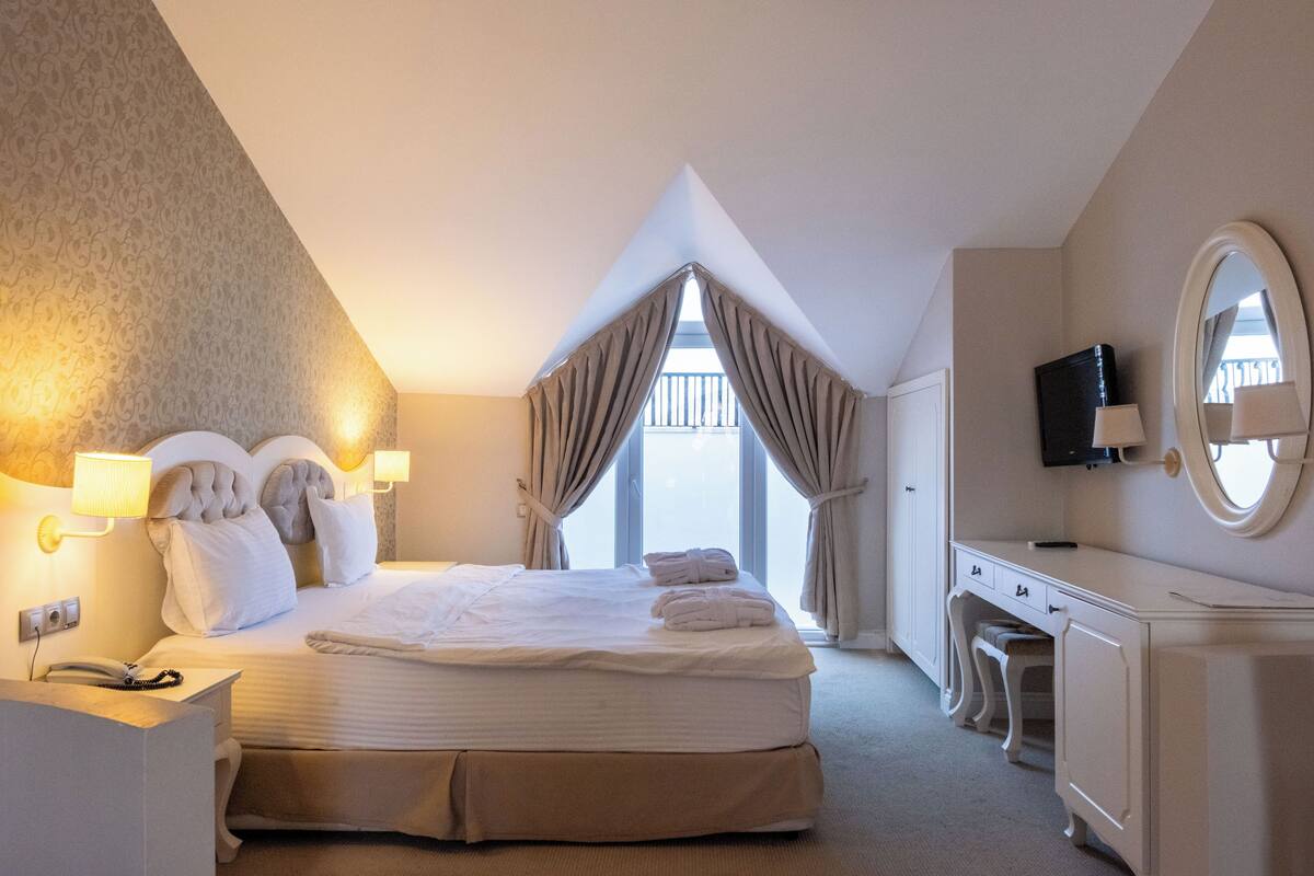A hotel room with a large bed, bedside lamps, a desk, a chair, a television, and a window with curtains.