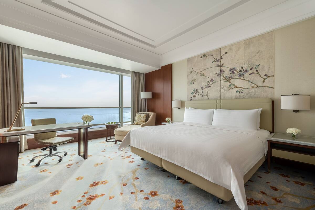 A hotel room with a large bed, a desk with a chair, a small table with a lamp, and a view of the sea through large windows.