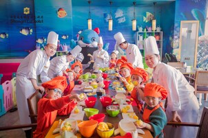 Un grupo de personas con gorros de chef alrededor de una mesa con comida y tazones coloridos, en una habitación con un revestimiento de pared temática de océano azul y repisas con utensilios de cocina.