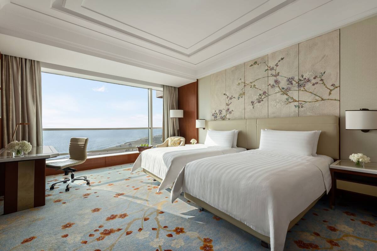 A hotel room with two beds, a desk, a chair, and a large window with a view.