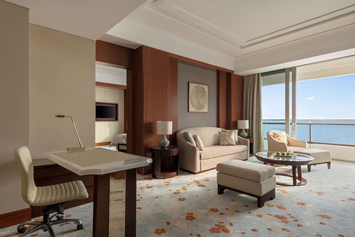 A hotel room with a large window, a sofa, a chair, a small table, and a desk.