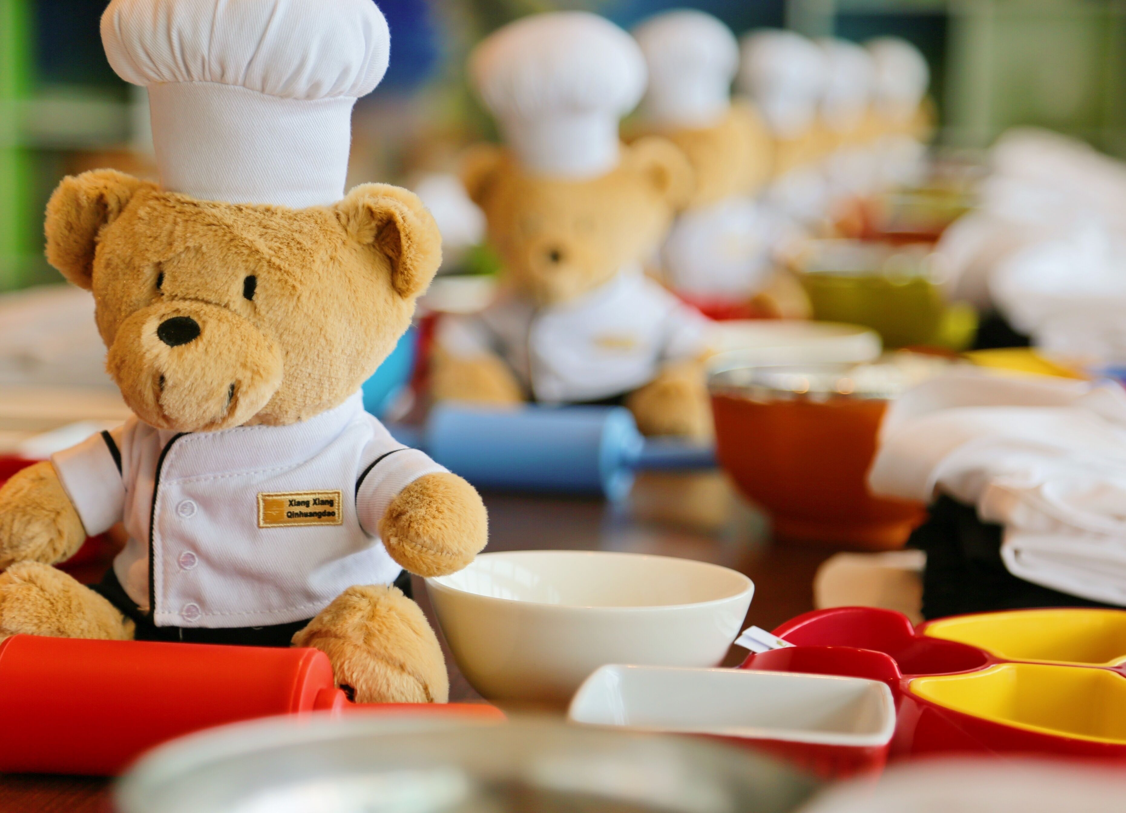 A teddy bear dressed as a chef with a name tag, surrounded by kitchen utensils and bowls.