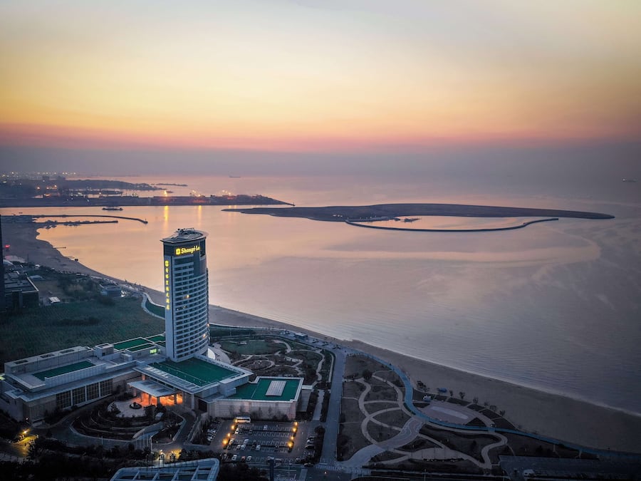 A high-rise hotel with a prominent tower, adjacent to a waterfront with a landscaped area and a parking lot.