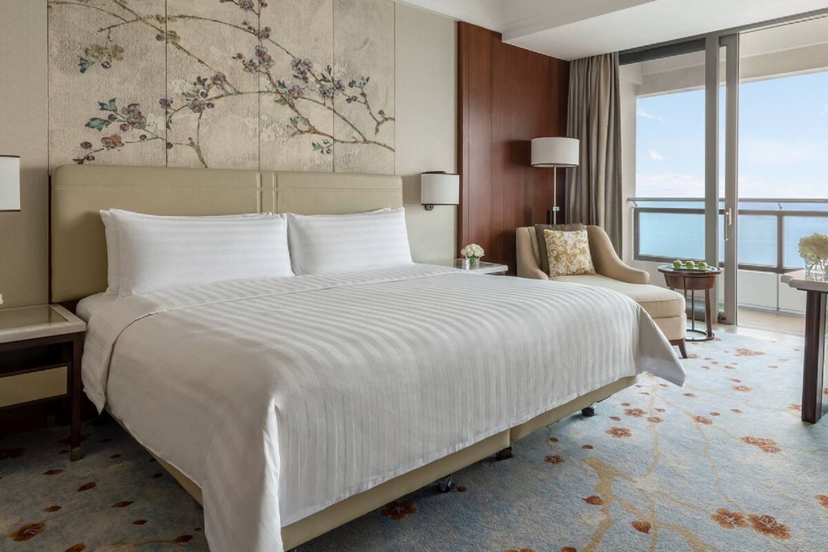 A hotel room with a large bed, two bedside tables, a chair, a small table, and a view of the sea.