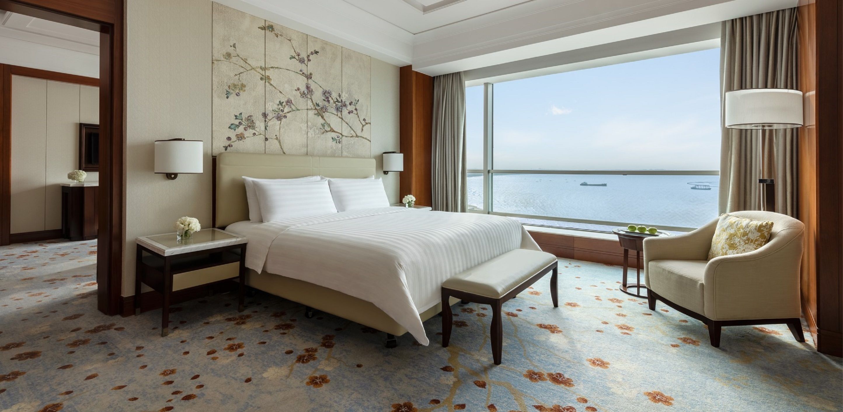A hotel room with a large bed, a sitting area with a chair, a nightstand, a bedside table, and a view of the sea.