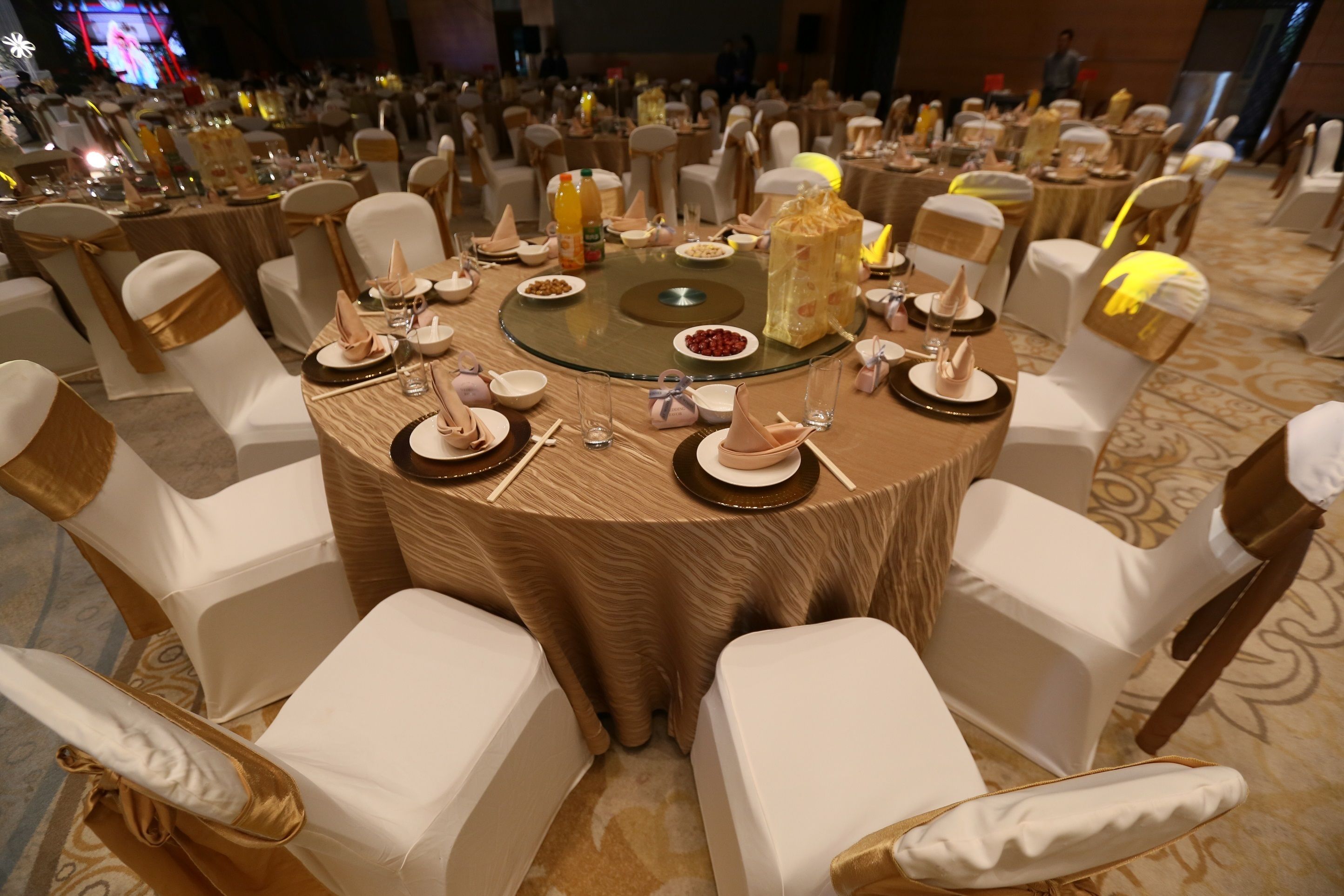 A round table set for a formal event with white chairs and gold sashes, surrounded by other similarly set tables.