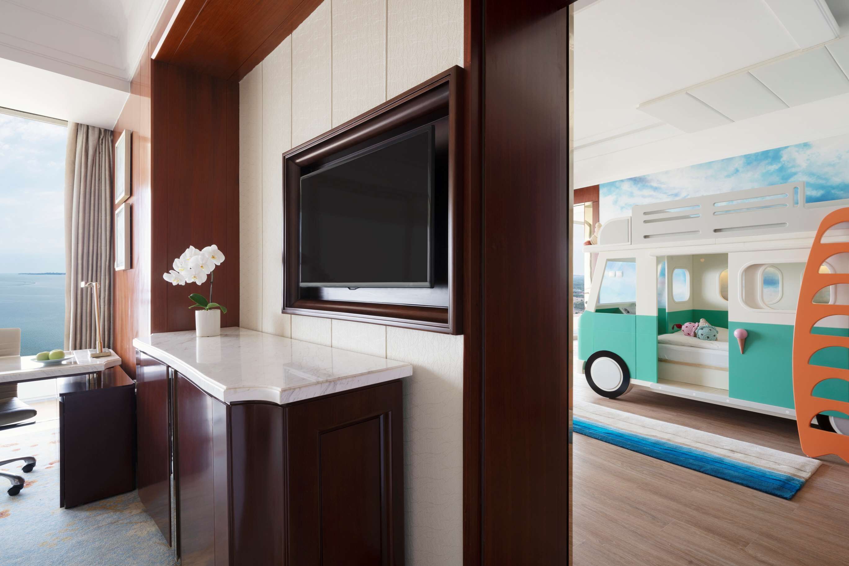 A hotel room with a large flat-screen TV mounted on the wall, a small desk with a chair, a potted plant, and a children's play area featuring a toy bus.