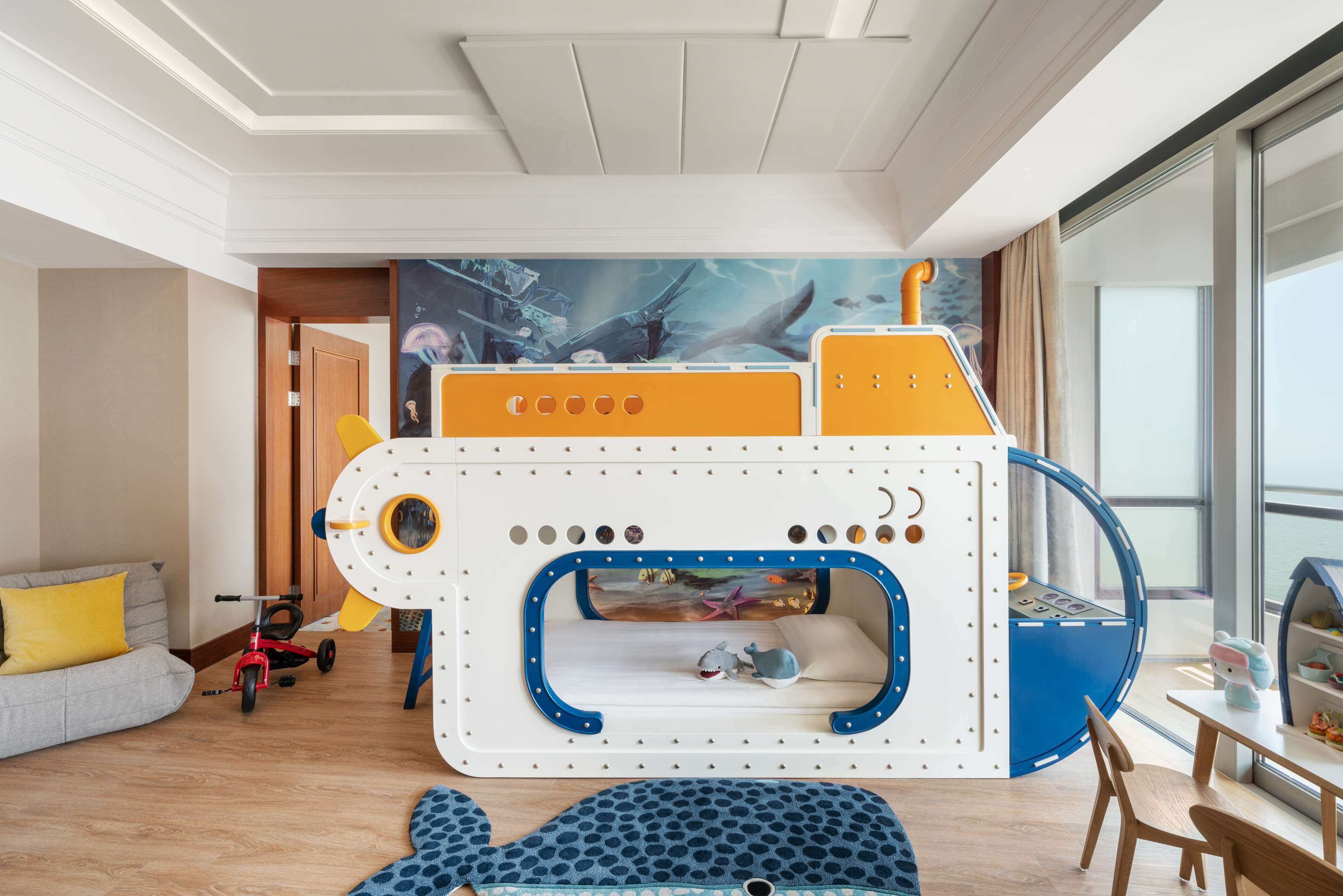 A child's play area with a ship-themed bed, a whale-shaped rug, and a window with a view of the sea.