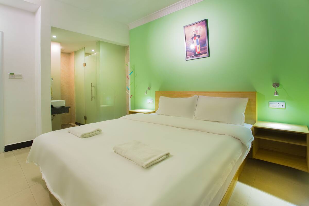 A hotel room with a large bed, a nightstand, a bedside table, a mirror, and a green wall with a framed picture.