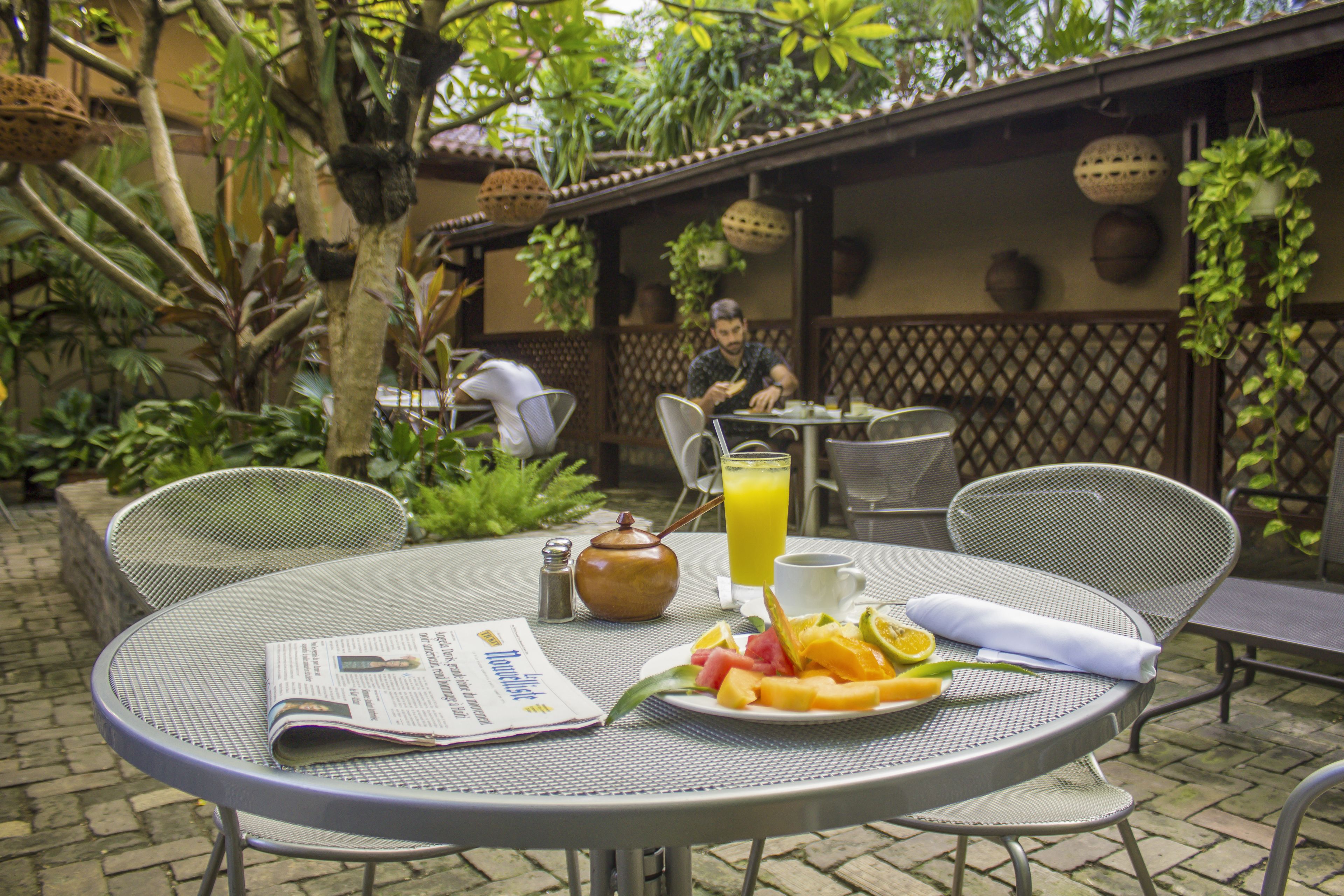 Un área de comedor al aire libre con mesas y sillas de metal, un plato de fruta, una tetera y un periódico.