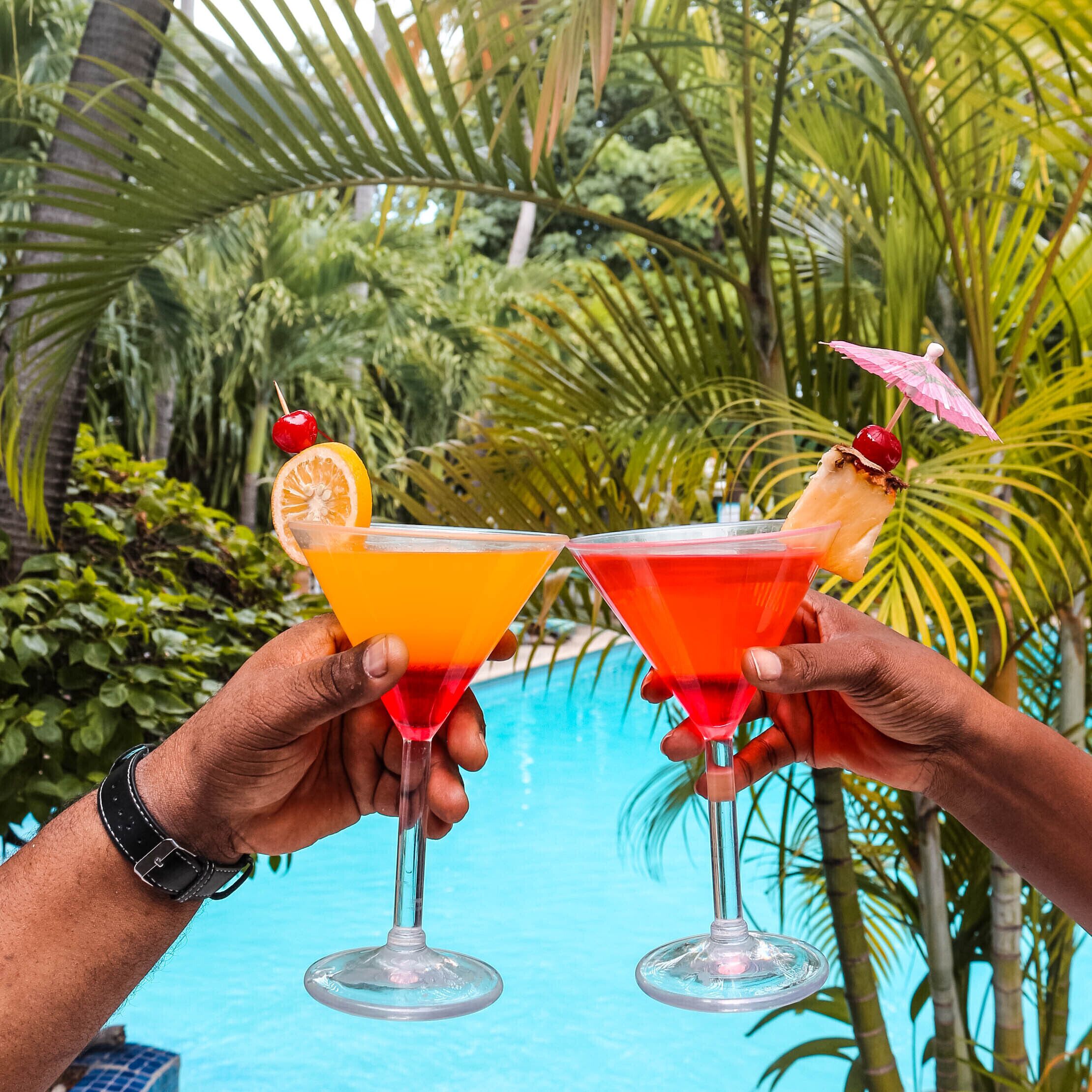 Two people holding martini glasses with tropical drinks and garnishes, standing by a pool with palm trees in the background.