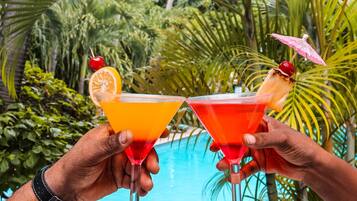 Two people holding martini glasses with tropical drinks and garnishes, standing by a pool with palm trees in the background.