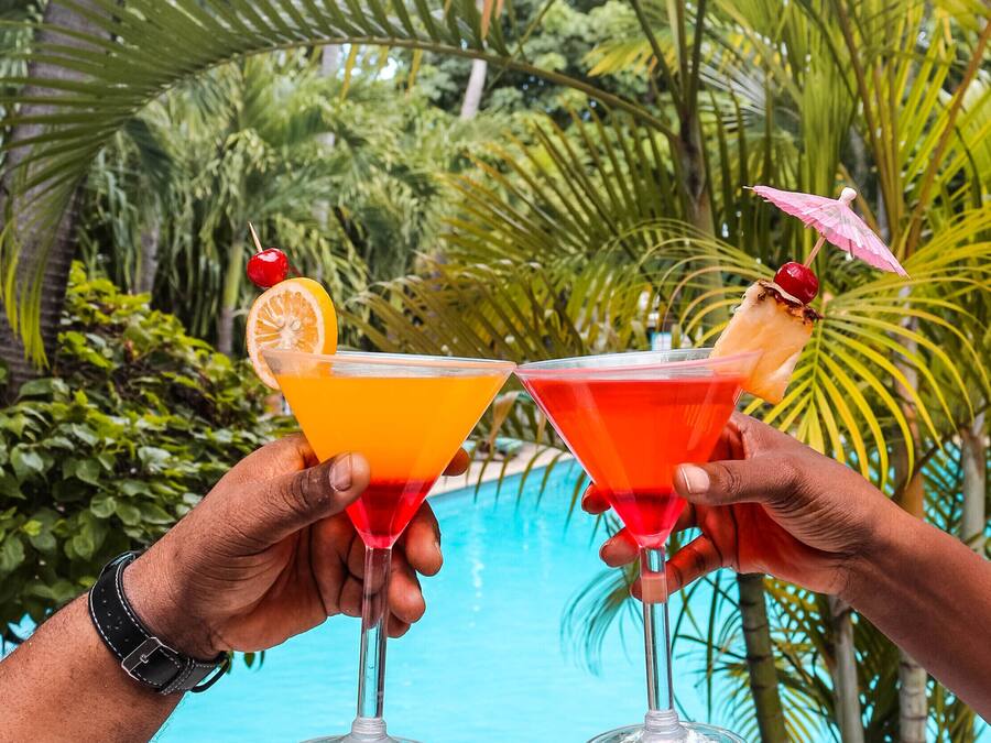 Two people holding martini glasses with tropical drinks and garnishes, standing by a pool with palm trees in the background.