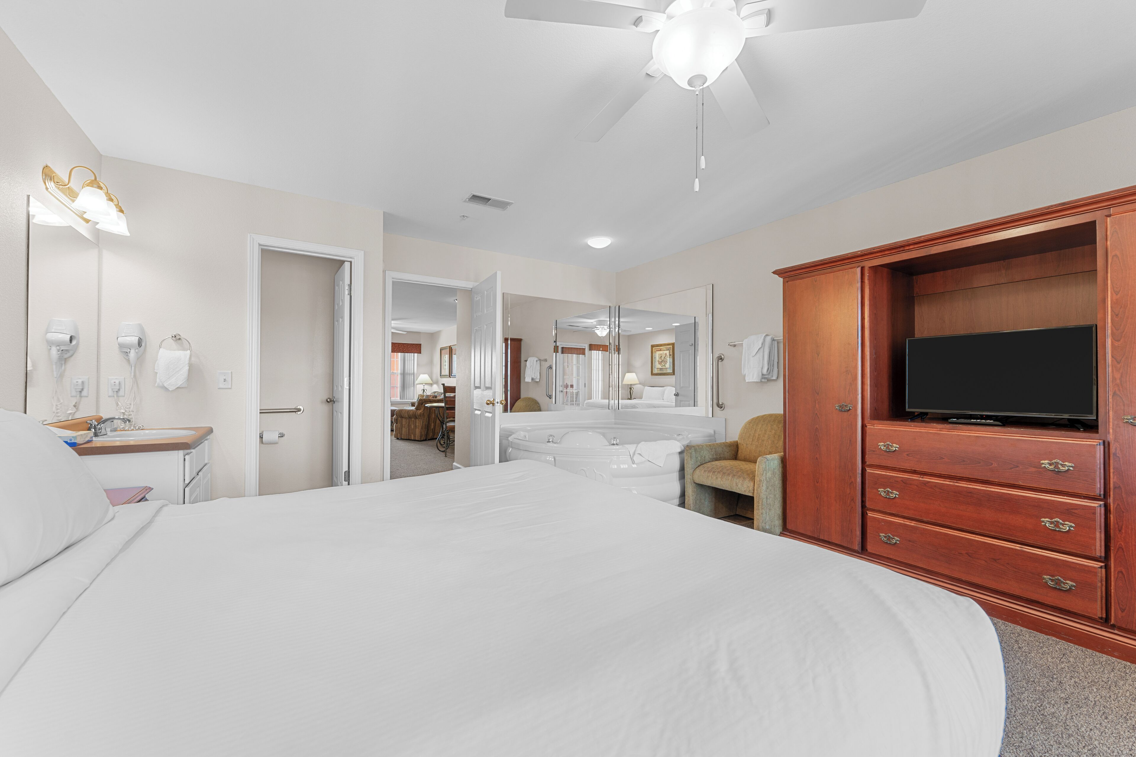 Une chambre d’hôtel avec un grand lit, une armoire en bois équipée d’un téléviseur à écran plat, un ventilateur de plafond et une salle de bain visible à travers une porte ouverte.