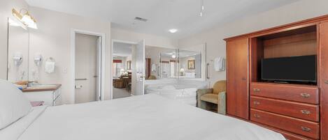 Une chambre d’hôtel avec un grand lit, une armoire en bois équipée d’un téléviseur à écran plat, un ventilateur de plafond et une salle de bain visible à travers une porte ouverte.