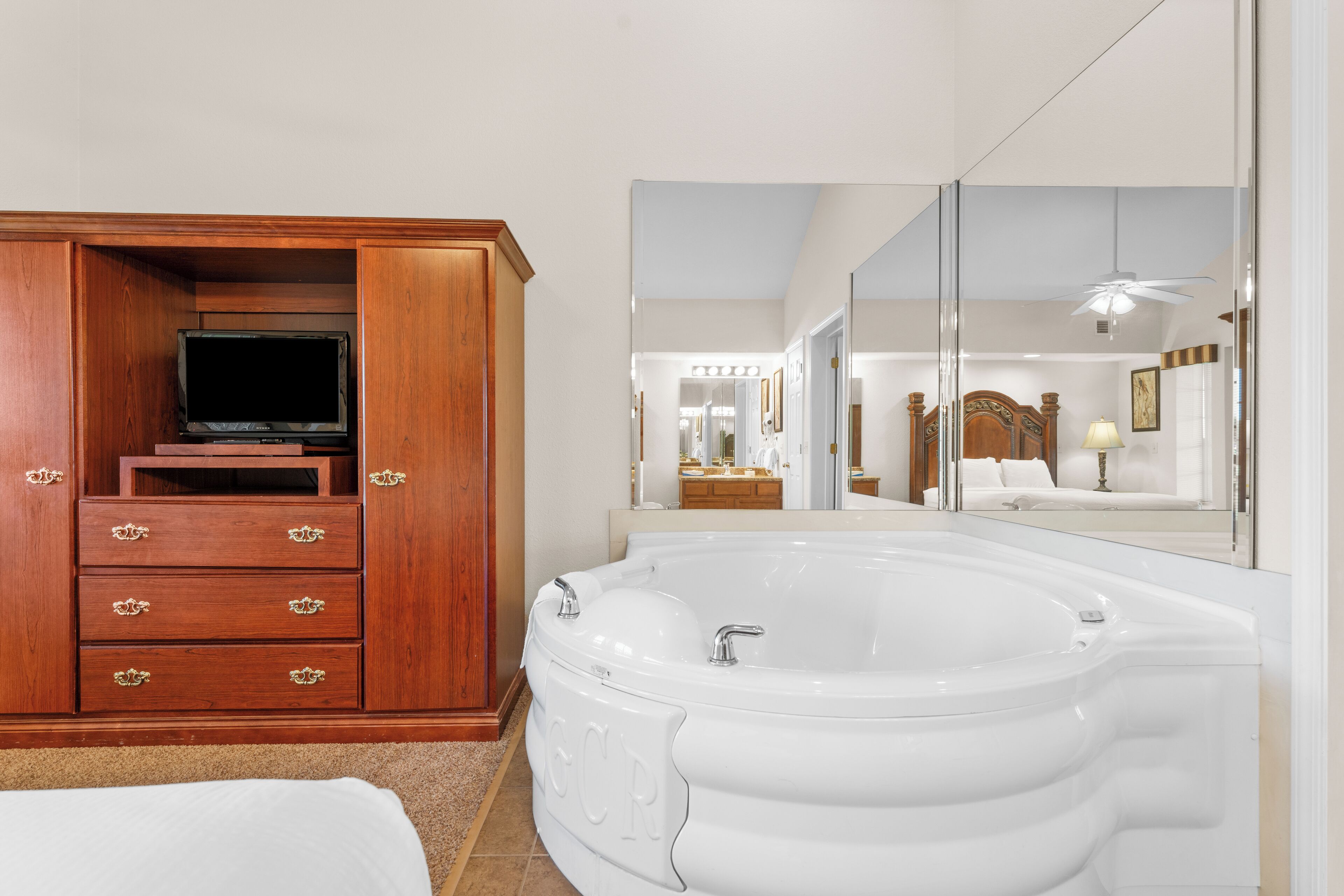 Une chambre avec une armoire en bois, un téléviseur à écran plat, une grande baignoire blanche et un miroir qui reflète la pièce.