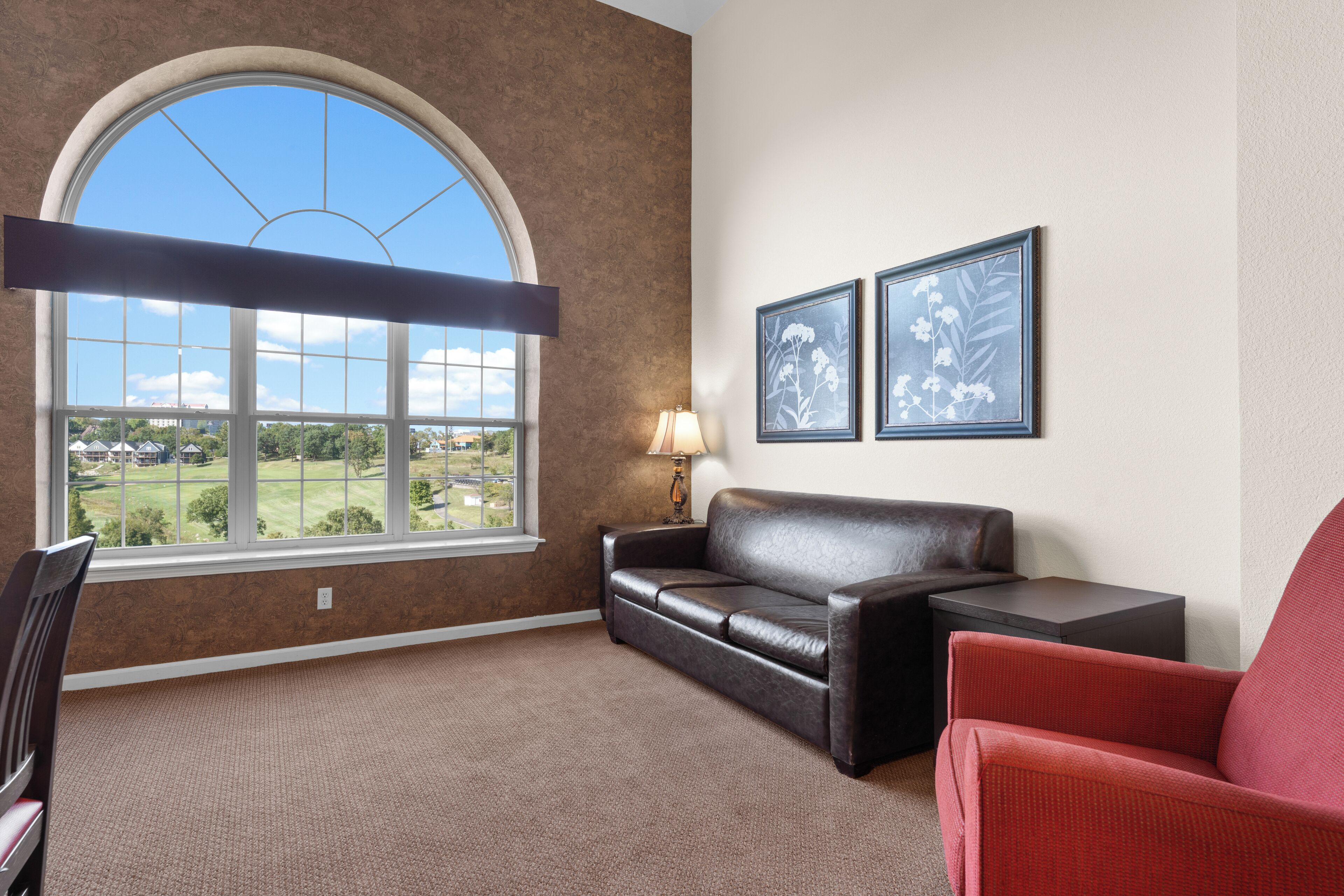 A room with a large arched window, a leather sofa, a red armchair, and two framed floral pictures on the wall.