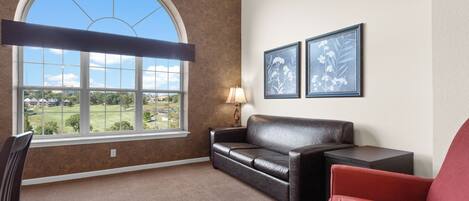 A room with a large arched window, a leather sofa, a red armchair, and two framed floral pictures on the wall.