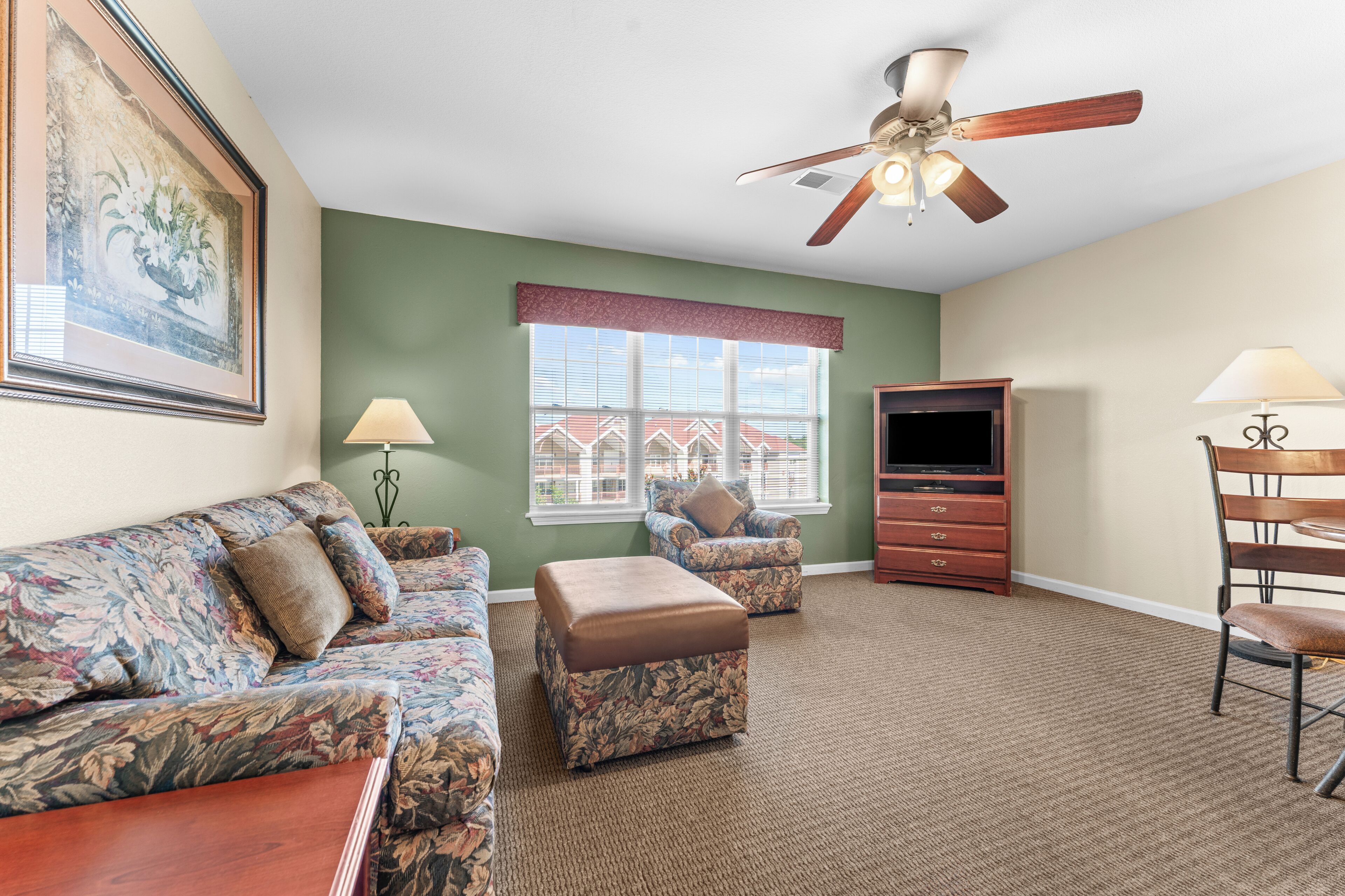 A living room with a green wall, a ceiling fan, a flat-screen TV, a wooden coffee table, and a floral sofa.