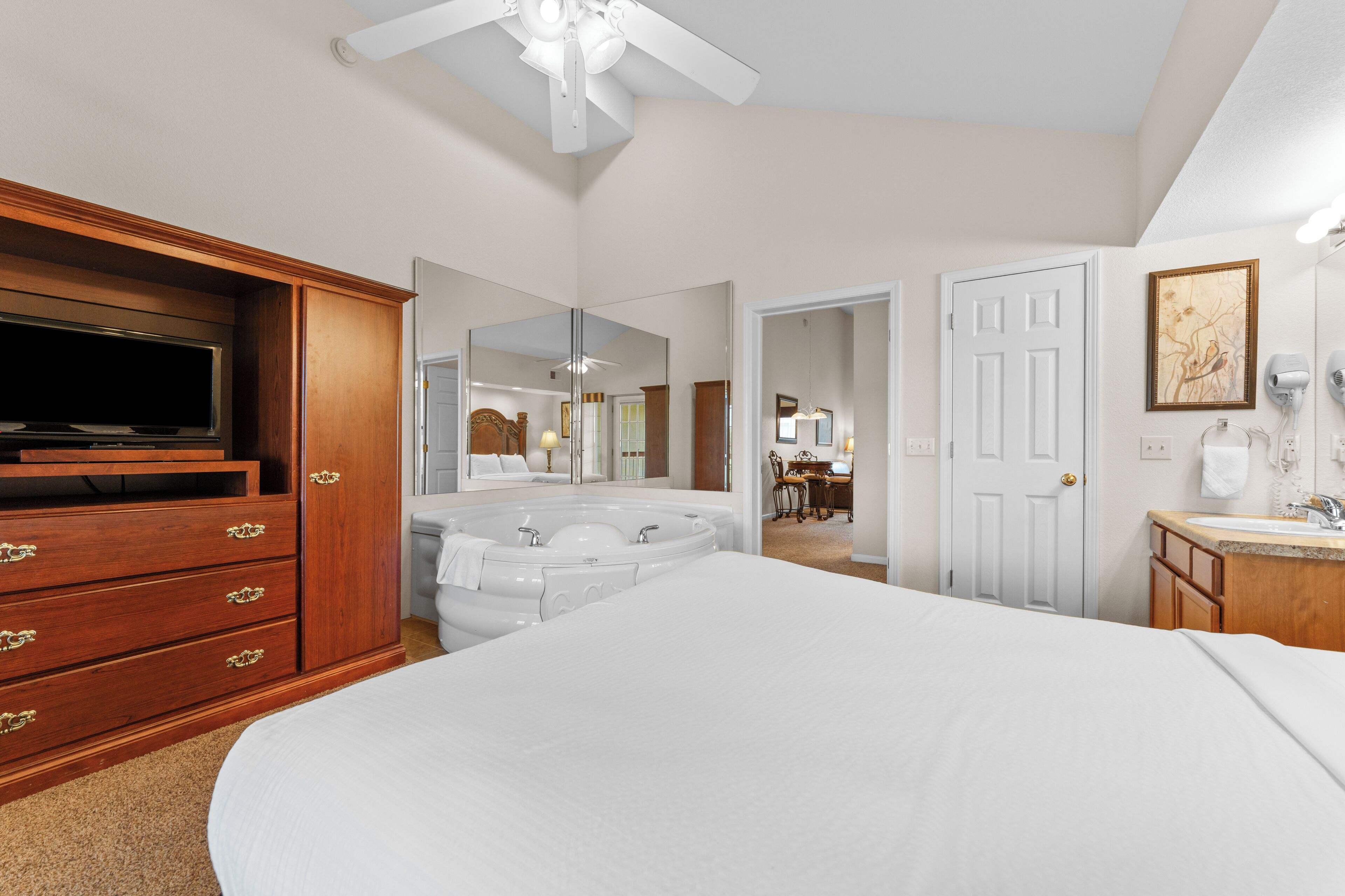 A spacious bedroom with a large bed, a wooden dresser with drawers, a TV, a white bathtub, and a bathroom with a sink and mirror.