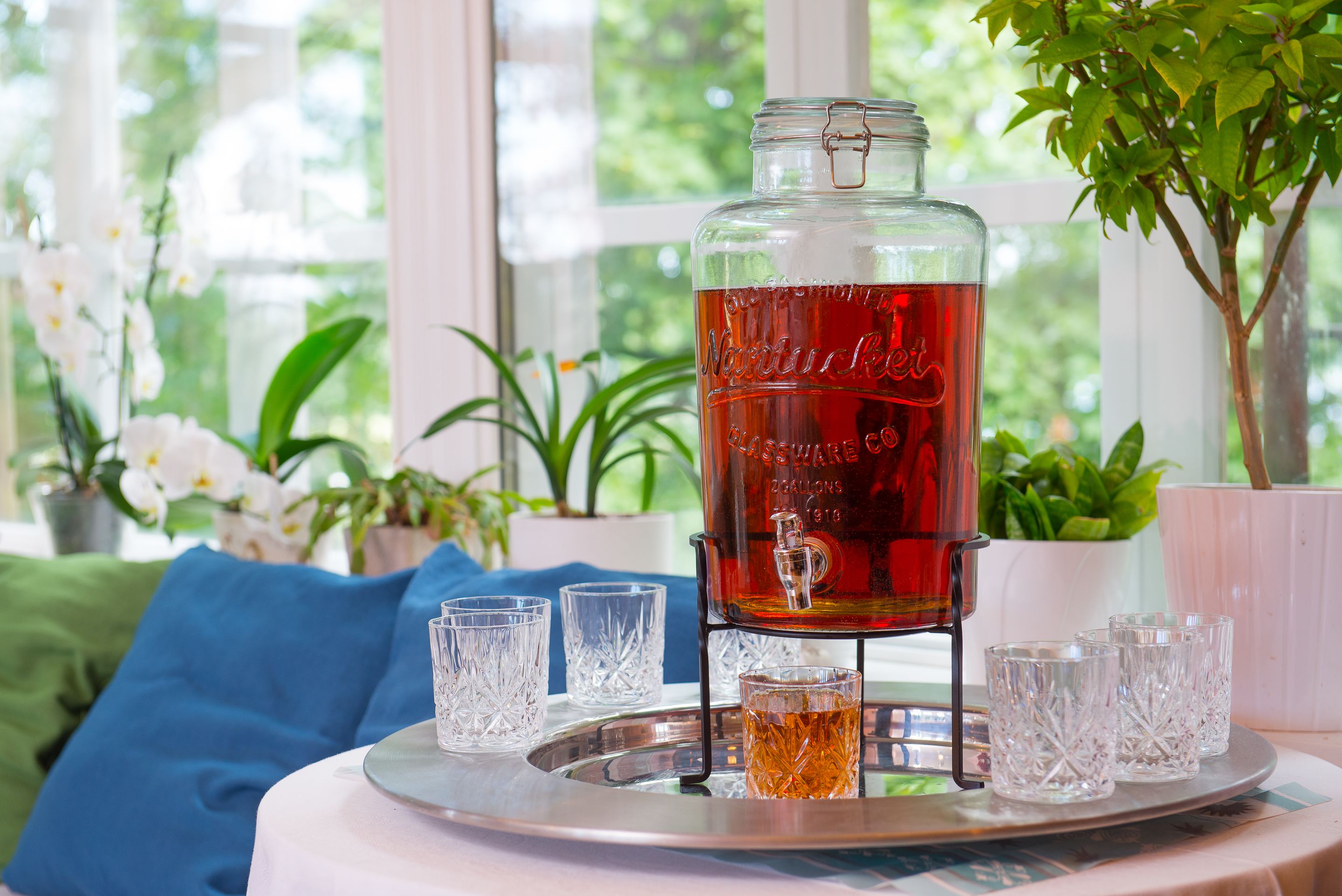 Una jarra de vidrio con una bebida roja sobre una bandeja con cuatro vasos, colocada sobre una mesa junto a una ventana con plantas.