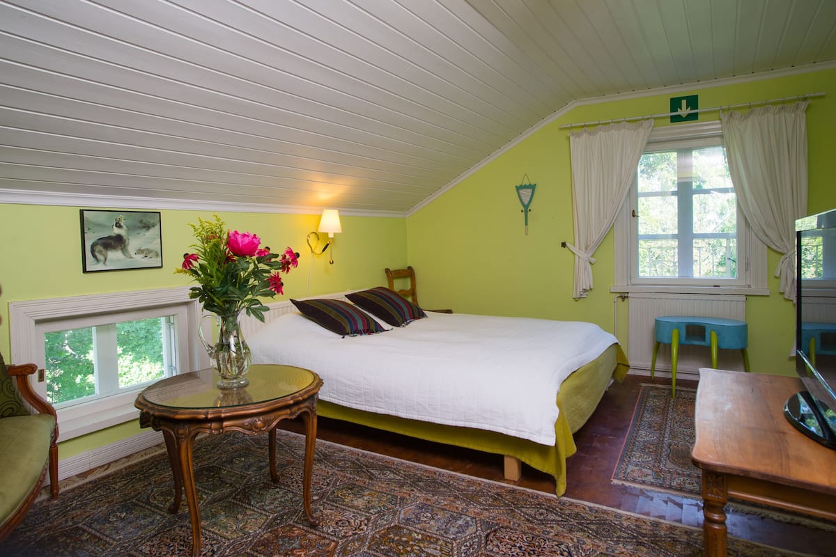 A bedroom with a bed, a table, a chair, a window with curtains, and a wall-mounted lamp.