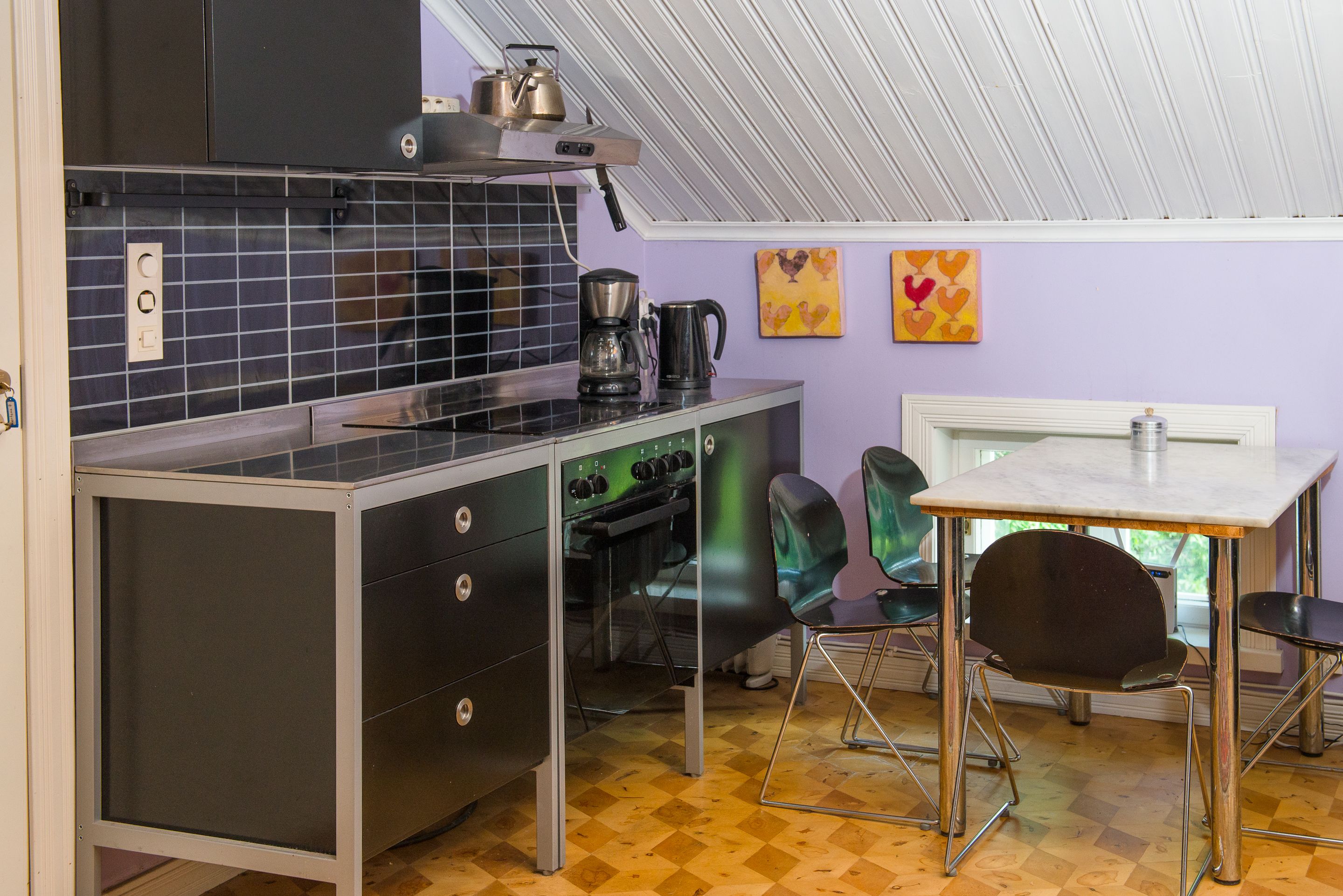 Een compacte keuken met een koffiezetapparaat, waterkoker en een klein oven. Er zijn twee stoelen en een tafel, boven een raam.