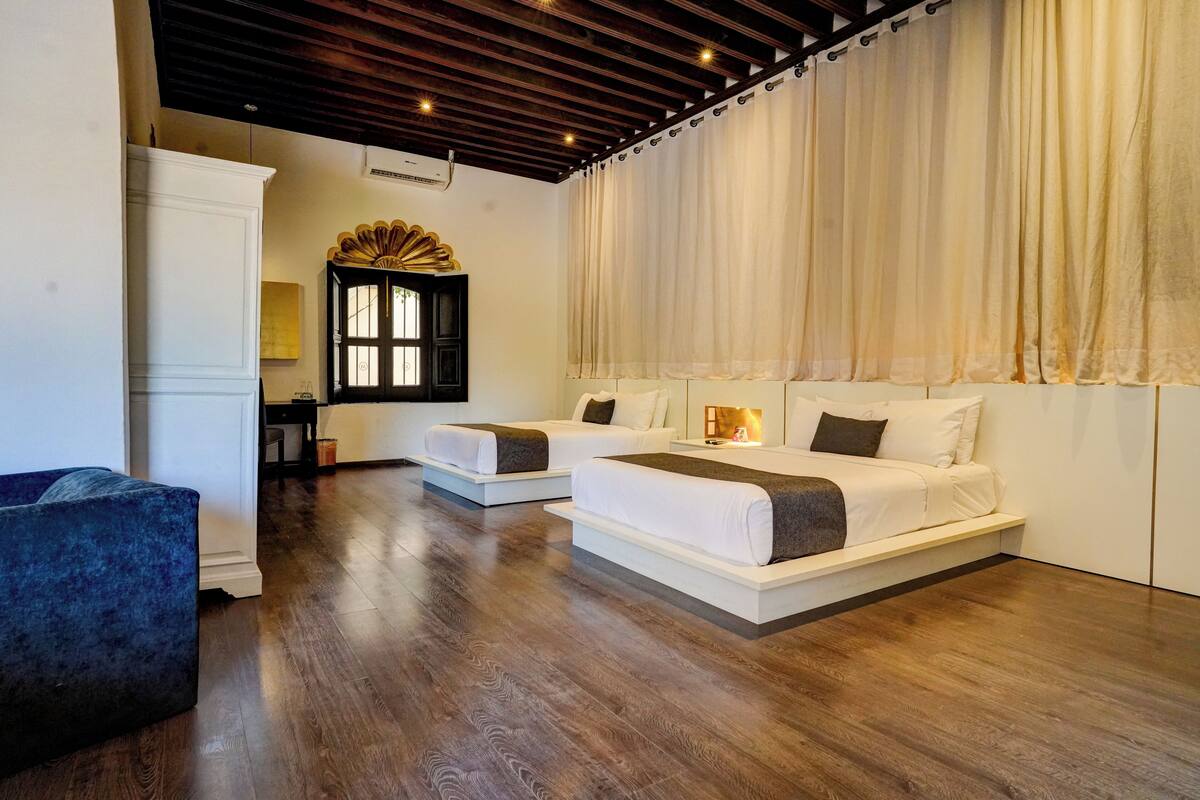 A hotel room with two beds, a blue armchair, a wooden ceiling, and a window with curtains.