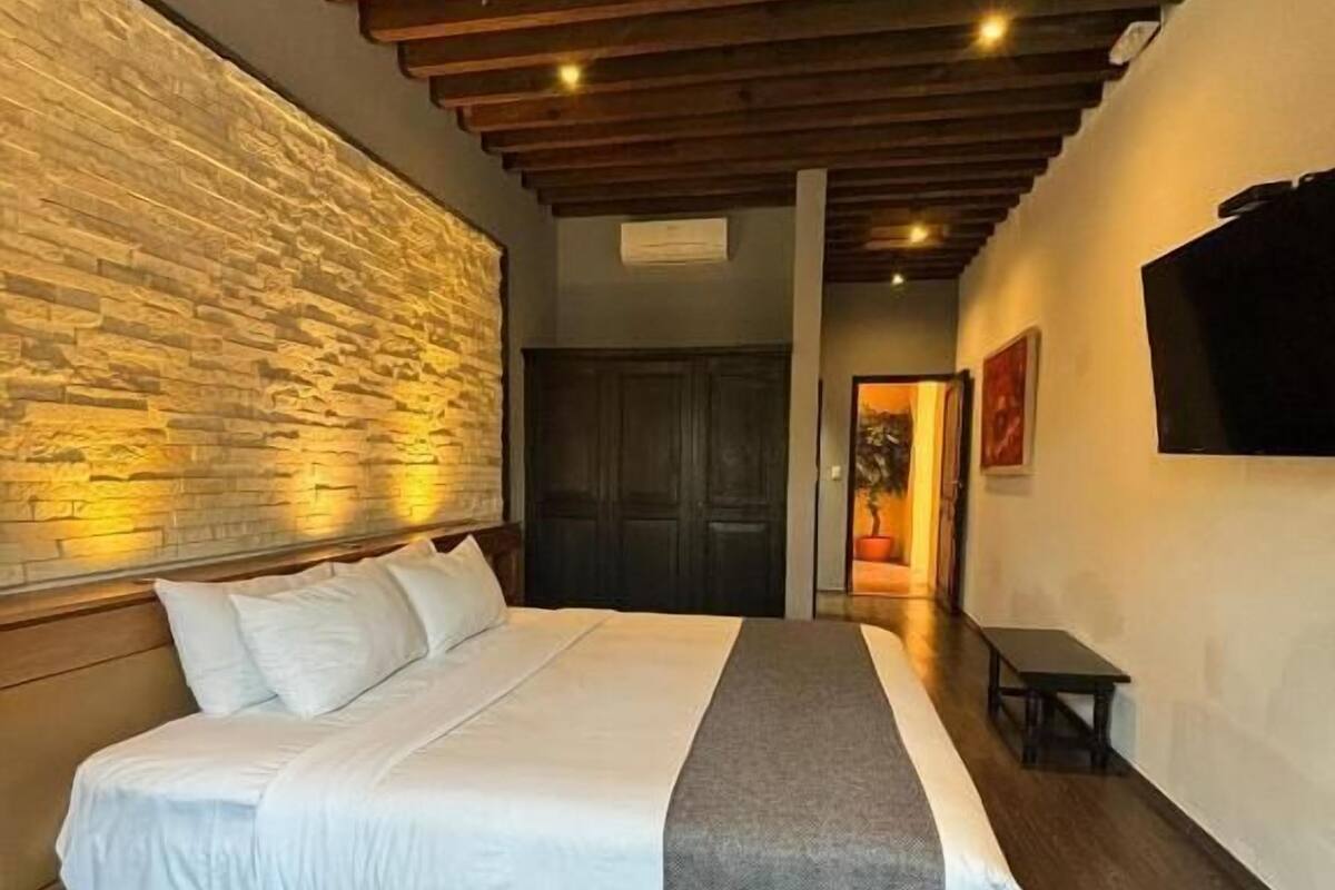 A modern bedroom with a large bed, wooden ceiling beams, a stone accent wall, and a flat-screen TV.