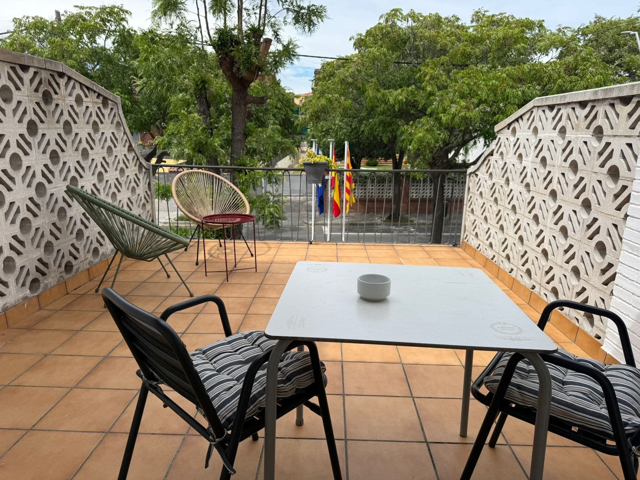 Un patio al aire libre con una mesa y sillas, un muro decorativo y vistas a árboles y edificios al fondo.