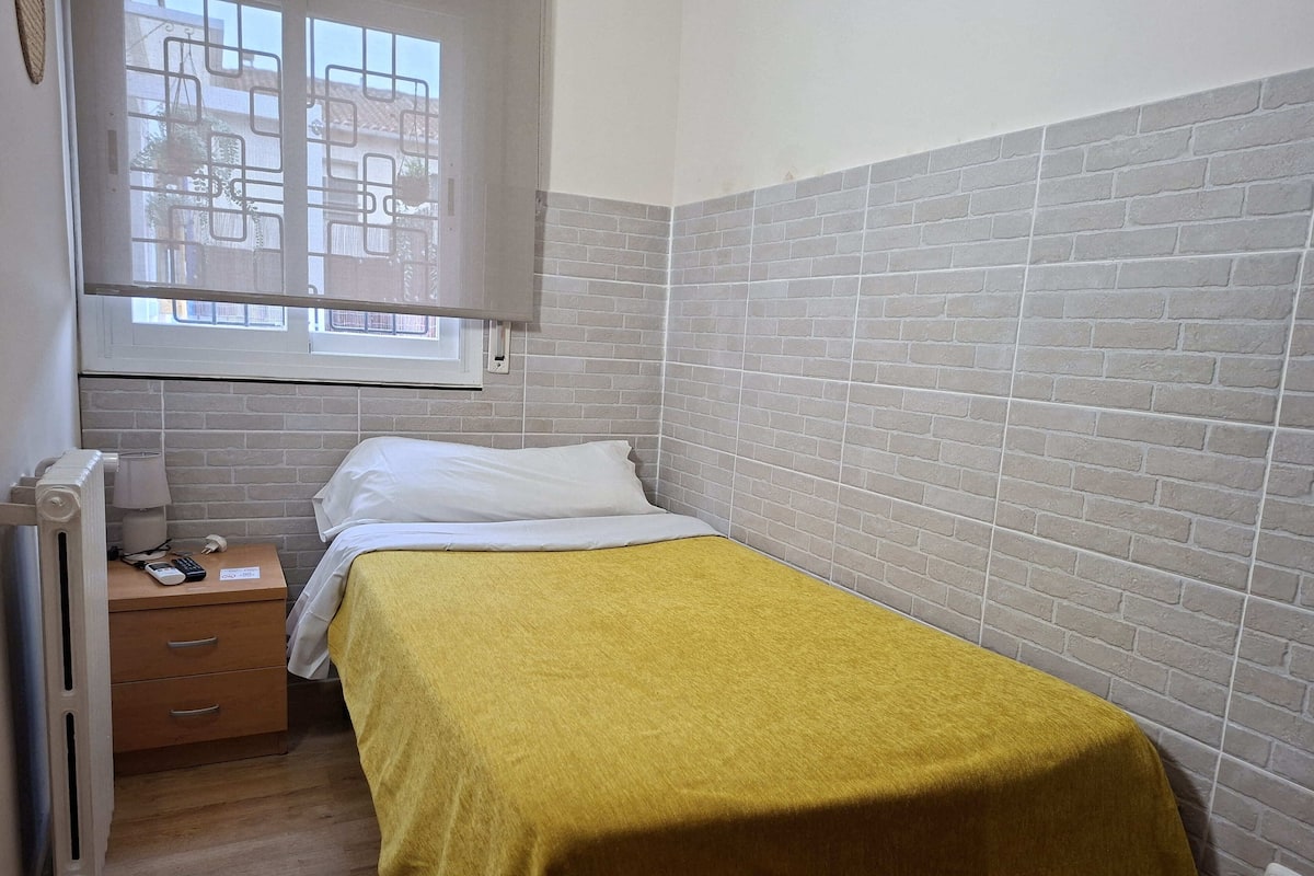 A small bedroom with a single bed, a nightstand, a radiator, and a window with a view of buildings.